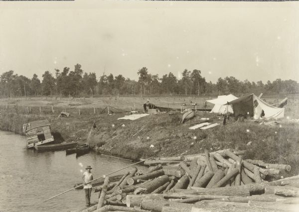 The travelers camp near the Northern Pacific Ridge. Canoes are docked at the edge of the river, and a pile of logs are in the foreground.