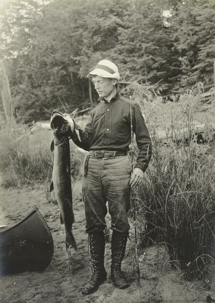 Former Senator Brazeau of Grand Rapids, Wisconsin posing near a canoe. He is holding his fishing pole and a large muskie.