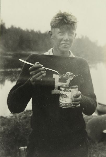 Russell Greene enjoying his dinner, which he is spooning from a can. He is standing by the river wearing a letter sweater.
