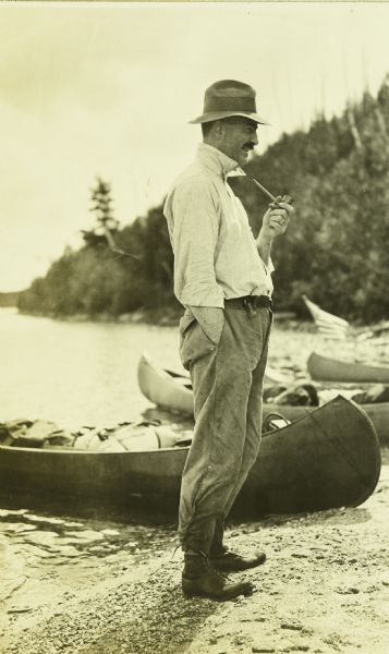 Bill Marr standing on the shore of the river next to canoes. He is holding a tobacco pipe.
