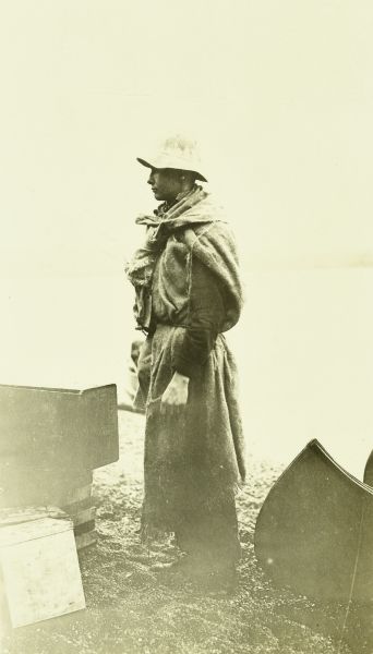 A young man wearing rain gear standing by a canoe and boxes of supplies.
