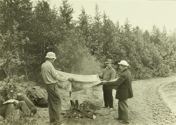 From L-R: Howard T. Greene, Bill Marr, and Doc Copeland holding a blanket over a campfire to dry it. The shoreline is on the right.