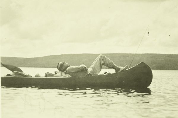 View across water towards one of The Gang reclining in a canoe on the water. His hat is pulled over his eyes and a fishing pole is propped up in the front of the canoe. The far shoreline is in the background.