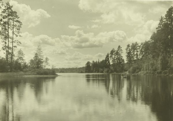 A view from the water of the French River and the tree-lined shorelines. There is a man in a canoe further down the river on the right.