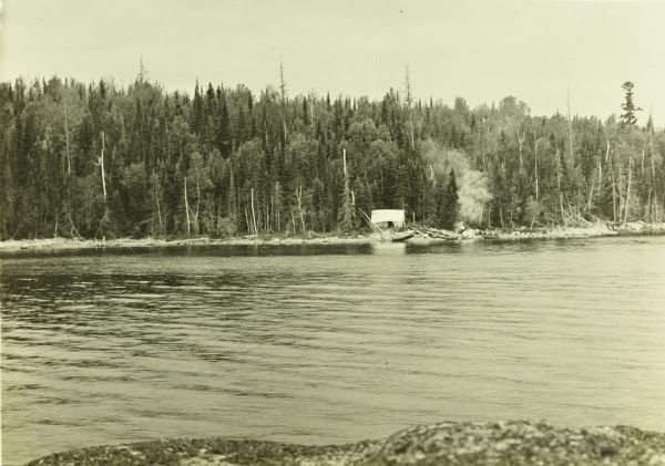 A view across Pickerel Lake of The Gang's camp on an island. There is a tent set up and smoke is wafting up from a campfire. 