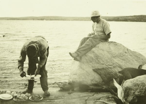 Two of The Gang eating a meal on the shore of Lake Pickerel. One of them is sitting on a large rock. There is a dog standing between rocks on the right.