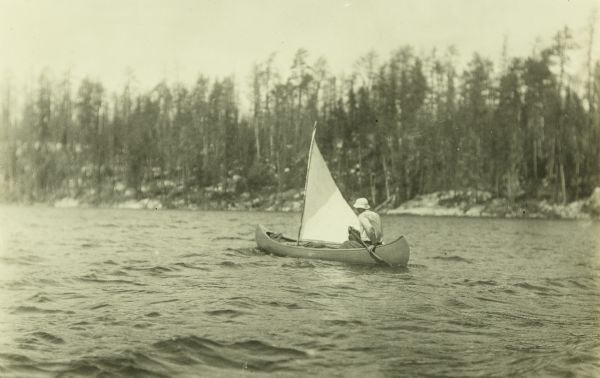 View across water towards Howard T. Greene traveling down a river in a canoe with a sail. The far shoreline is in the background.