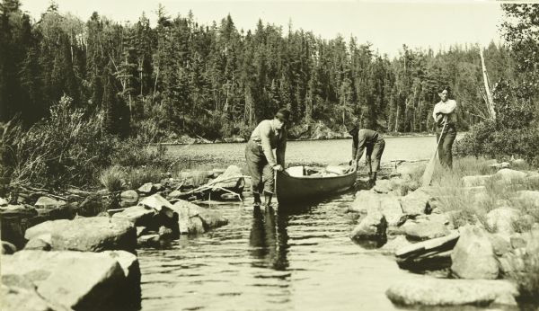 The Gang lifting their canoes through a narrow, shallow passage into Batchewaung Lake. The passage is lined with rocks. One of The Gang is leaning on a paddle and standing on the rocks.