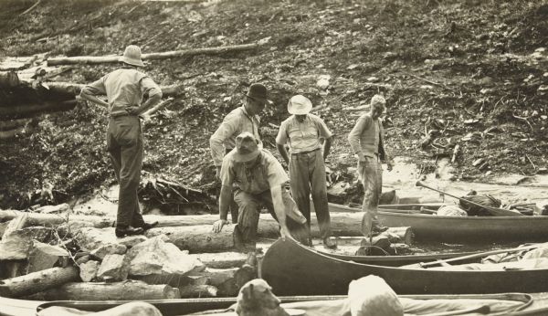 Five of The Gang standing among rocks and logs in the shallow portion of the Quetico River. They are standing next to their canoes, which are loaded with supplies.