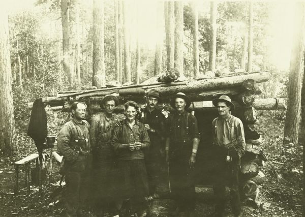 An outdoor group portrait of the Canadian Rangers at their camp. Their names are Wellington "Wallie" Musselman, Fred Hampshire, Robert Johnson, Stanley Wall, Addison Cox, and George Wall (brother of Stanley). The men, all of whom are from Ontario, are posing in front of a log structure.