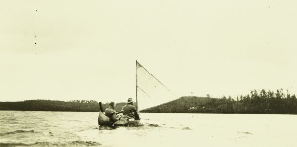 View across water towards two of The Gang (possibly Carl and Bill) gliding on Rouge Lake in a canoe with a sail. The far shoreline is in the background.