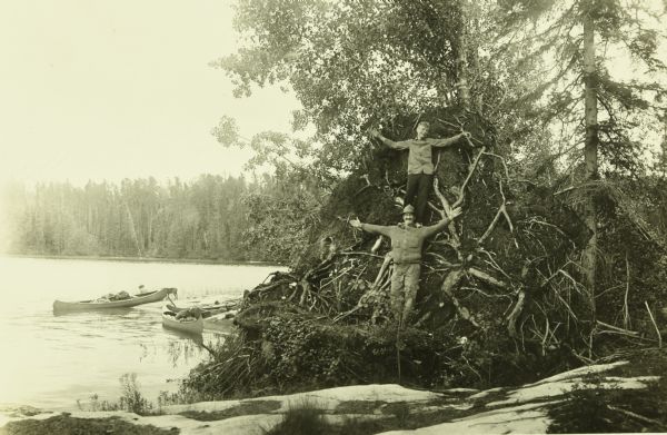 Carl Greene and Bill Marr posing and standing with their arms spread wide at the base of a large, uprooted tree at the shoreline. Their canoes are in the water of Jean Lake on the left. 