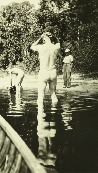 Carl, and one of The Gang standing with a dog, are bathing in Sturgeon Lake. Another man is standing on the shore.