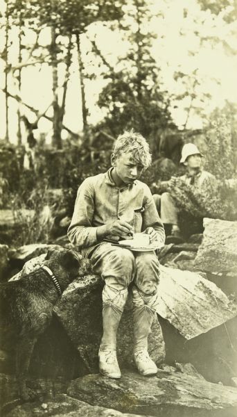 Carl is sitting on a rock with a plate of food on his lap. His dog Diadem (Di) is at his side. One of the men is sitting behind them.
