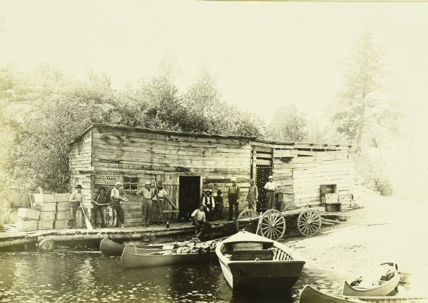 View across water towards the Gang standing in front of G. Swenson's Store in Kettle Falls, which is partially on the water. The steamer schedule is handwritten on left side of front of building. A boat and canoes are at the shore and there is a wagon parked near the store.