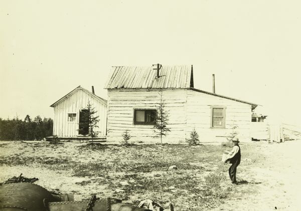 "Old Bill," a Native American, is carrying a basket or box outdoors in Kettle Falls. There is a large wooden building behind him.