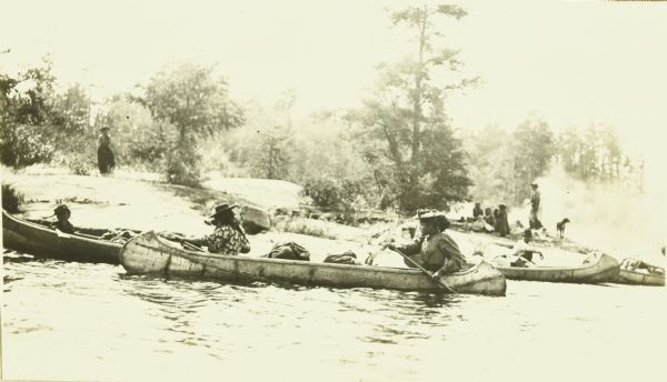 Indian women paddle their canoes on Rainy Lake. Other people and dogs are on the shore.