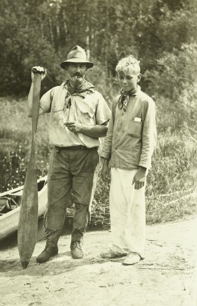 Bill Marr and Carl standing together at the end of the Dawson Trail trip. Bill is holding a paddle, and a canoe is behind them on the left.