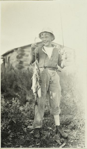 Jack Green proudly holding up several fish he caught. There is a log cabin in the background.