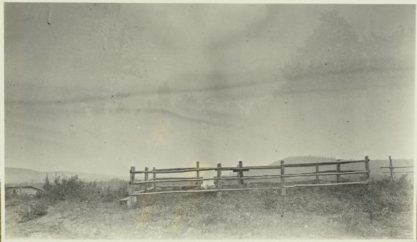 View towards a wooden fence surrounding a cemetery near Tom Lake, north of Chicago Bay. Wooded hills are in the background.