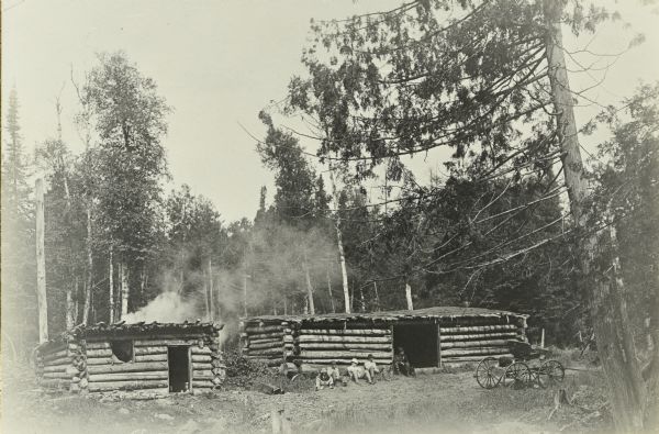 Several members of The Gang resting outside the log cabins at Half Way Camp on Tom Lake, north of Chicago Bay. There is a carriage on the right.