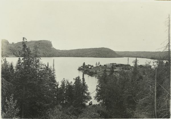 Elevated view looking over trees towards the lumber camp on McFarland Lake, which was owned by the Pigeon River Lumber Company.