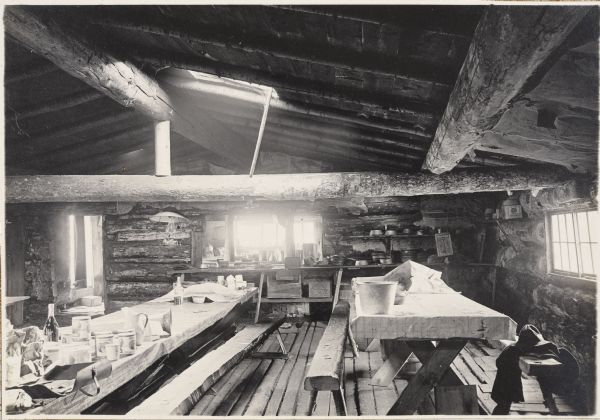 An interior view of the kitchen at the Pigeon River Lumber Camp at  McFarland Lake. There are cooking utensils and supplies on long tables and counters. The walls and ceiling of the shack are made of logs, and there is a skylight near the center of the roof.