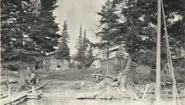 View across water towards a log cabin and an outbuilding on the homestead of Edwin Kugler between Pine and McFarland Lakes, north of Chicago Bay. There is a pier on the left.