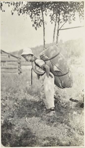 Jack carrying a large pack on his back. There is a log cabin in the background.