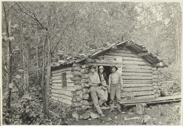 From left to right are Clay, Jack, and Edwin Kugler posing in front of Kugler's trapper's shack near his homestead at the end of Pine Lake. Edwin has a pipe in his mouth.