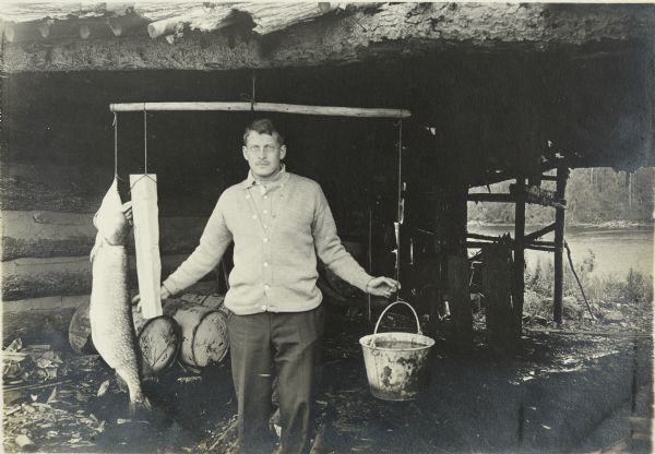 Mackey weighing a large fish near a log cabin. The lake is partially visible in the background on the right. 