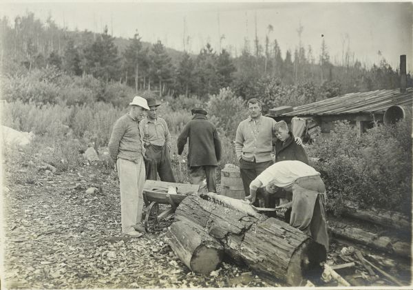 The Gang is watching as McGrew is scaling and gutting a large fish on a log outdoors. Doc Copeland is standing on the left wearing a light-colored hat. Mackey Wells has his arm around Jack. The other two men are unidentified.