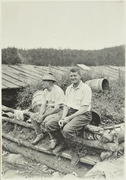 Doc Copeland (left) and McGrew sitting together on a fence made of logs. There is a building with a long roof and a skylight in the background.
