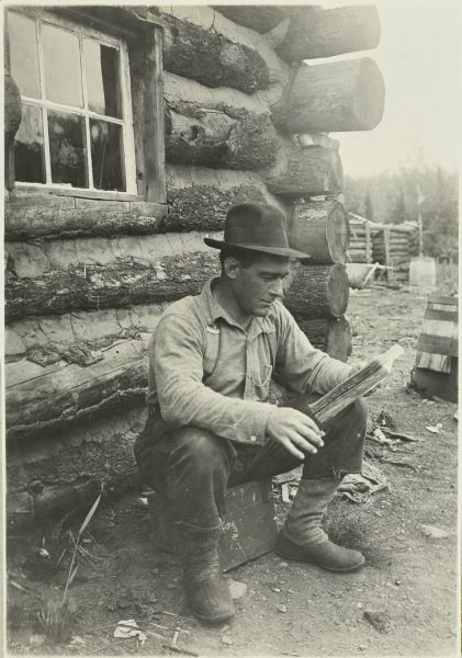 Mullen sitting next to a log cabin and reading at a lumber camp on McFarland Lake.