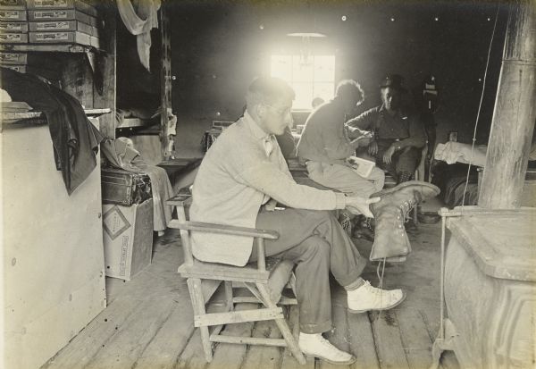 Mackey Wells sitting and holding a boot. Two men are sitting behind him; one of them has reading material on his lap. There is a woodburning stove in the foreground on the right.