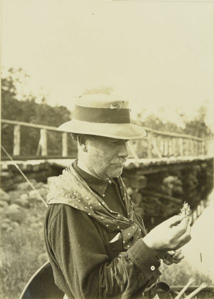 Billy Mac examining a plant. He is wearing a hat and a kerchief. There is a bridge behind him.