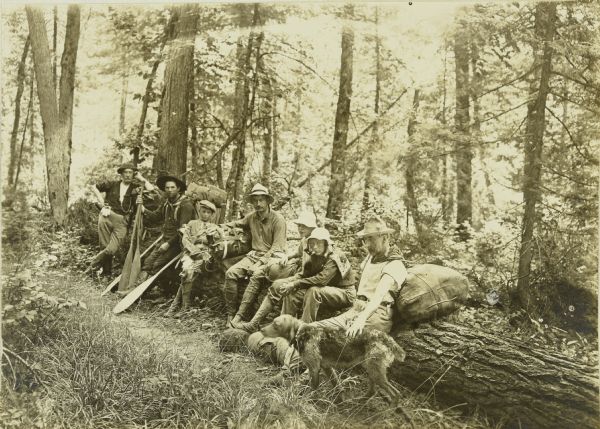 The Gang resting on a fallen tree during their first portage of the trip. From left to right are: Billy Mac, Fred Carr, Carl, Bill Marr, Howard T., Charles, Doc Copeland, and Nimrod the dog. Several of them are holding canoe paddles.