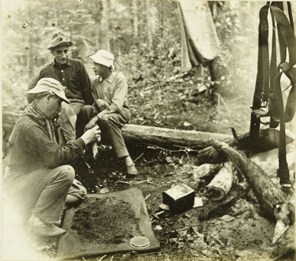 Billy Mac spreading tobacco on a board to dry as Doc Copeland is watching from his seat on a log. Carl is sitting next to Doc.