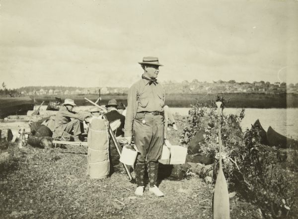 William MacLaren, known to The Gang as Billy Mac, standing with camping supplies including a bedroll, a jug, and a box. A few of his travel mates are sitting behind him.