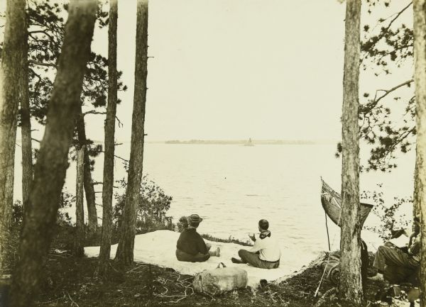 Billy Mac and Dad sitting on a tarp, looking out over the lake. Another person is sitting on the right. There is a flag displayed that may be an Ontario flag.