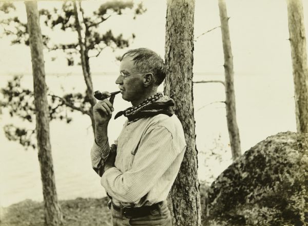 Howard "Dad" Greene looking contemplative as he is smoking his tobacco pipe outdoors. There is a lake in the background.