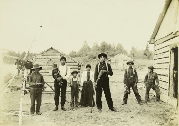 John Otter, who is standing second from the left, posing with seven members of his family in an Indian village at the mouth of the Namakan River. There is a birch frame on the left which is used for drying jerky.