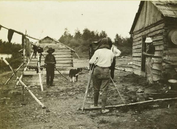 A view of Howard "Dad" Greene photographing an Indian man and a dog next to a jerky-drying frame. They are in an Indian village at the mouth of the Namakan River.