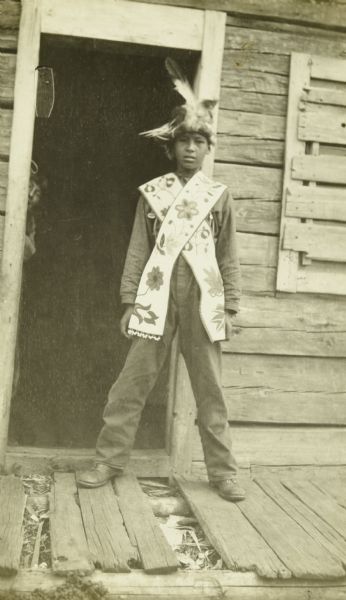 An Indian boy posing wearing a feathered headdress and a beaded bandolier in front of the doorway of a building. He is in an Indian village at the mouth of the Namakan River.