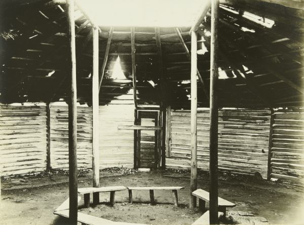 An interior view of a dance house at an Indian Village. The house has an open roof and benches inside.