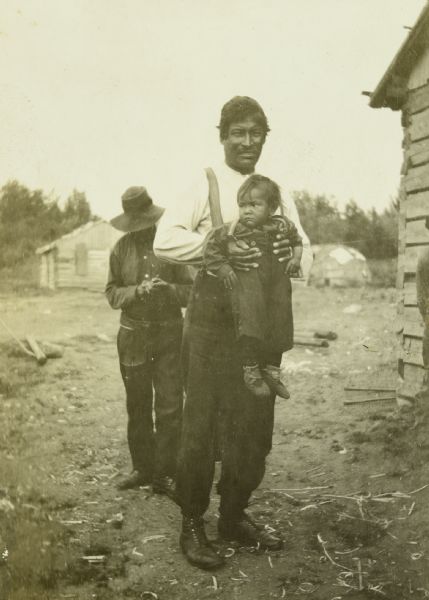 An Indian man is standing and holding a small child in his hands in an Indian village by Namakan Lake. Another man is standing behind them, and a wikiup is in the background.