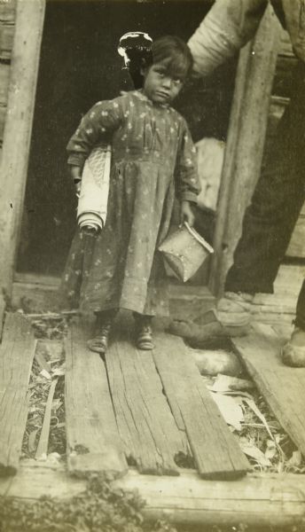 A young Indian girl standing on a run-down porch. She is wearing a dress and is holding a small basket in one hand, and a rolled item under her other arm. There is an adult, partially obscured, standing next to her on the right.