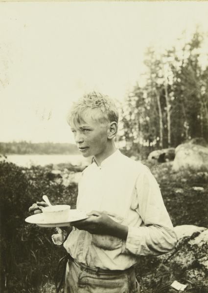 Carl standing outdoors eating food from a plate he is holding. There is a lake in the background beyond rocks and trees.
