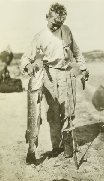Carl posing outdoors holding a very large fish, with a fishing pole leaning against his shoulder.