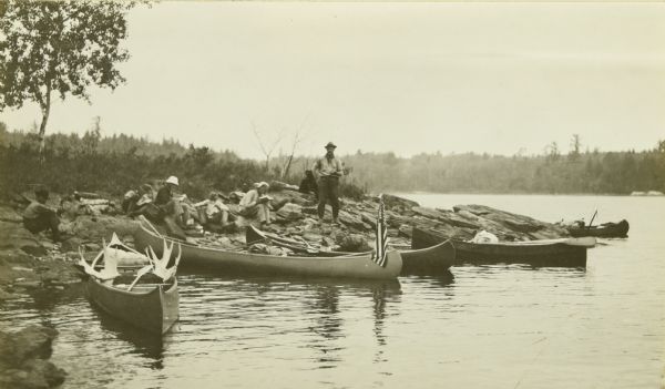 The Gang stopping at Sand Point for a lunch break. The canoes are at the edge of the shoreline, while everyone is sitting on the ground eating. The canoe on the left is decorated with moose antlers, and another canoe displays an American flag.